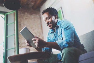 Bearded smiling African man using tablet for video conversation while relaxing on sofa in modern office.Concept of young business people working at home.Blurred background.Selective focus.