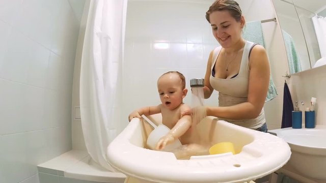 Young smiling mother washing her baby son in bathtub from showerhead