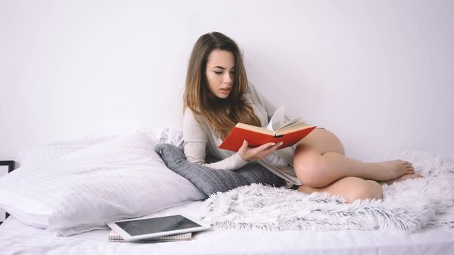 Young smiling woman reading book while lying in bed in her bedroom