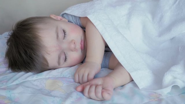 Baby boy 2 years old sleeping in a crib covered white blanket.