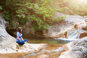 Fototapeta na wymiar Young woman meditating in lotus position while doing yoga in a wonderful forest near waterfall. Travel, Healthy Lifestyle concept