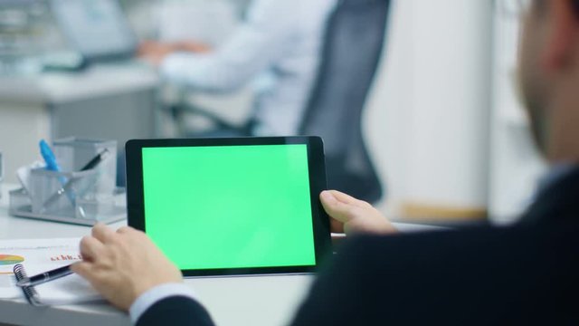 Businessman Uses Tablet Computer with Green Screen, He Swipes and Touches Screen. In the Background His Colleague Works on a Laptop. Office is Bright and Modern. Shot on RED Cinema Camera 4K (UHD).