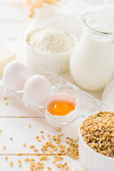 Ingredients for baking: flour, milk, wheat grain, butter and eggs on white wooden background. Selective focus
