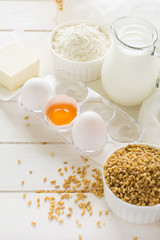Ingredients for baking: flour, milk, wheat grain, butter and eggs on white wooden background. Selective focus
