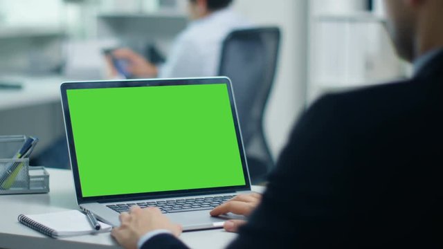 Businessman Working on a Laptop with Green Screen on. In the Background His Colleague Working on a Tablet Computer. Office is Modern and Bright. Shot on RED Cinema Camera 4K (UHD).
