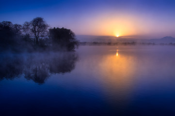 霧に煙る湖面に映る日の出と浮島