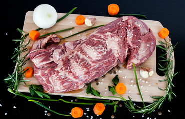 Big piece of raw beef meat with vegetables onion, carrot, garlic and spices pepper, rosemary, parsley, sea salt on a wooden cutting board.
