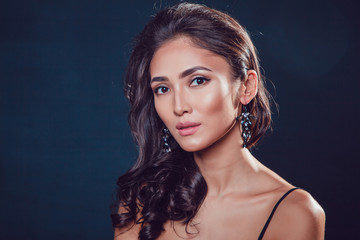 Brunette girl with shiny wavy hair and beautiful earrings posing at studio on black background.