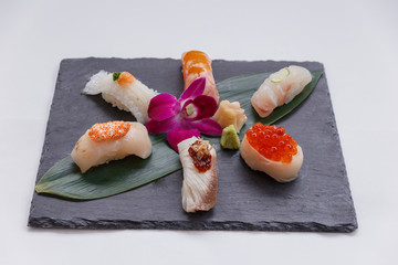 Sushi Set Include Torched Salmon, Engawa, Hotate, Hamachi, Tai and Ikura with Hirame Served with Wasabi and Prickled Ginger on Leaf on Black Stone Plate.