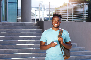 smiling african student walking by stairs with bag and smart phone