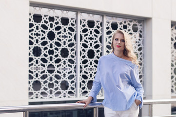 Beautiful european female with long blond hair stand in near white wall with arabic pattern. Young business woman in blue blouse and white trousers