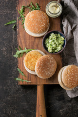 Ingredients for making vegan burger. Veggie cheese and onion cutlets, yogurt sauce, hamburger buns, herbs and avocado salad, served on wooden board with textile on dark background. Top view