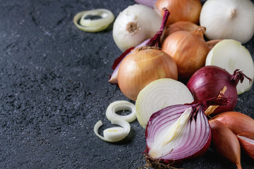 Variety of whole and sliced red, white, yellow and shallot onions over dark stone texture background. Close up, space for text