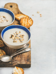 Healthy dinner with creamy mushroom soup in ceramic bowls and grilled bread slices on rustic serving board over grey marble background, selective focus, copy space