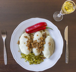 Potato dumplings - a traditional regional dish. Polish and Lithuanian cuisine. With pickles , fried bacon and paprika . White plate, knife, fork and glass of vodka on a dark background..