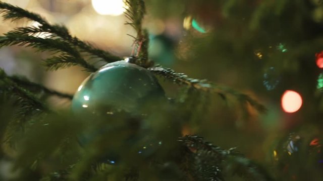 Close up shot of green christmas ball hanging on a fir tree branch against background with colorful bokeh lights.