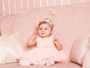 Smiling baby girl 1 year old wearing trendy pink dress and floral hairband sitting on sofa in room.