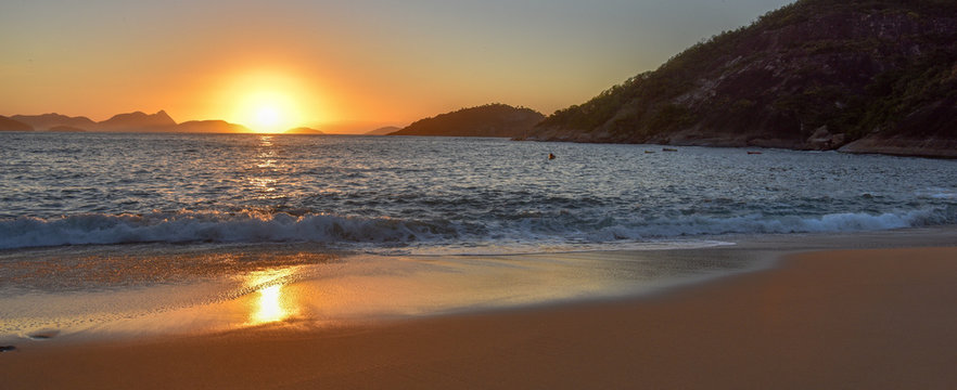Beautiful sunrise with the sun rising from the Atlantic Ocean, solar path on the water and sand at the deserted Praia Vermelha Beach, Rio de Janeiro, Brazil