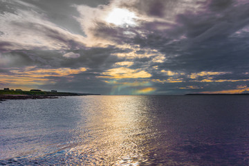Orkneys, Scotland - June 5, 2012: Seascape looking at entrance to silver to mauve Atlantic Ocean under dark cloudy evening sky wherein waning sun creates colorful lights. 