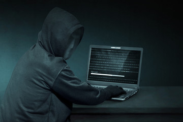 Hooded hacker with anonymous mask using laptop to steal data