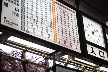 A display board and a clock in its side can be seen in this image where the display is sharing some information. Few trees leaves can be seen behind the display.