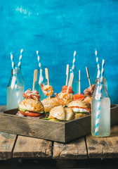 Home party food concept. Homemade burgers with sticks in wooden tray and lemonade in bottles with straws on rustic shabby table, bright blue painted wall at backgound, selective focus, copy space