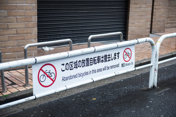 A road blocking iron railing is seen on the picture. The iron railing is marked with no parking symbols and written in Japanese language. On the background, a building with shutter is seen.