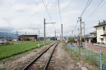 In the picture we can see a railway road , field and some buildings. The railway overhead wire can also be seen in the picture.