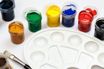 Open gouache jars with empty white plastic palette and few paintbrushes on a white background