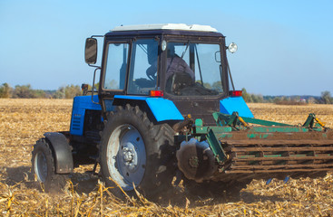 Fototapeta premium Big blue tractor plows the field and removes the remains of previously mown corn. Part of the cultivator, steel, round discs in a row close-up. Work agricultural machines. Harvest.