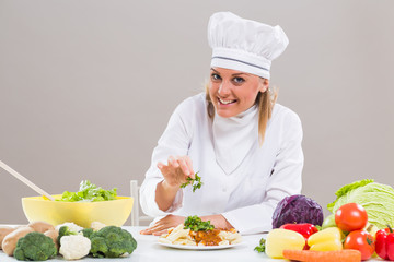 Cheerful female chef is sitting at the table with bunch of vegetable and decorating prepared meal.
