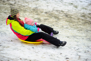 Fototapeta na wymiar Young woman with little daughter together move down from a hill on saucer sled.