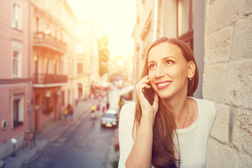Young beautiful woman standing on the balcony and speaking on cell phone on street of european city. Mobile communication copy space image