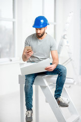 Handsome repairman or builder in helmet working with drawings and phone on the renovation of apartment interior