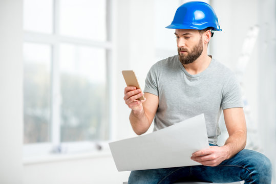 Handsome repairman or builder in helmet working with drawings and phone on the renovation of apartment interior