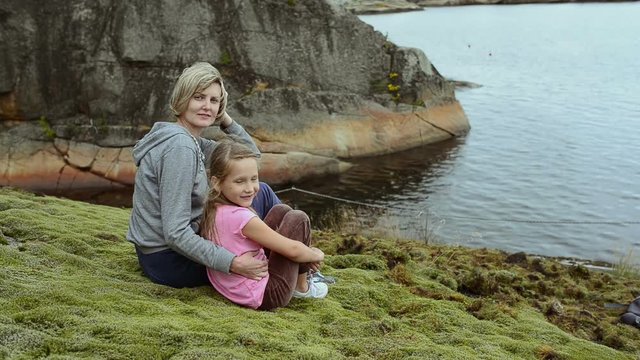 Mother and daughter sitting on the big stones on the sea shore, looking at camera