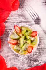 Strawberry salad, a kiwi and orange against a dark background with icing sugar and red fabric
