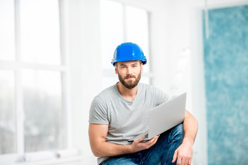 Portrait of a handsome builder, foreman or repairman in the helmet sitting with laptop in the white...