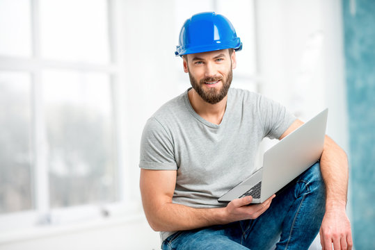 Portrait of a handsome builder, foreman or repairman in the helmet sitting with laptop in the white interior