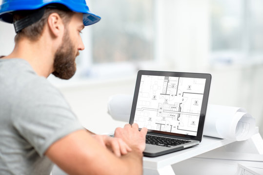 Handsome architect or foreman in helmet working with laptop on the apartment drawings at the structure white interior
