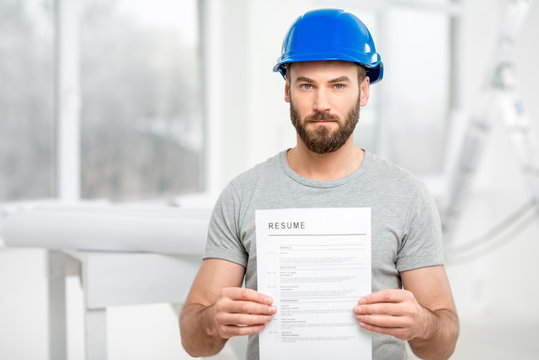 Handsome builder or worker in protective helmet holding his resume standing in the white interior.
