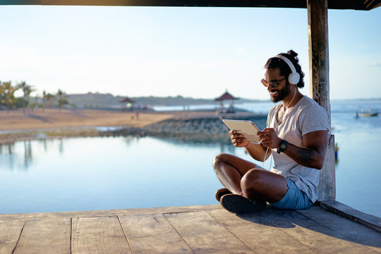 Relaxed and cheerful. Work and vacation. Outdoor portrait of happy young african man using tablet computer on deck near the sea.