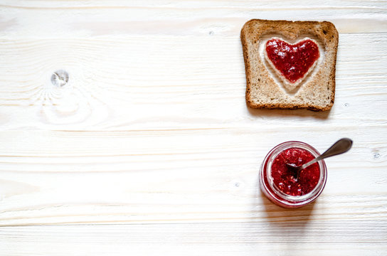 Toasted white bread with a heart inside. At the heart plastered with raspberry jam. Under the croutons is a jar of jam, a jar of sticks dessert spoon.