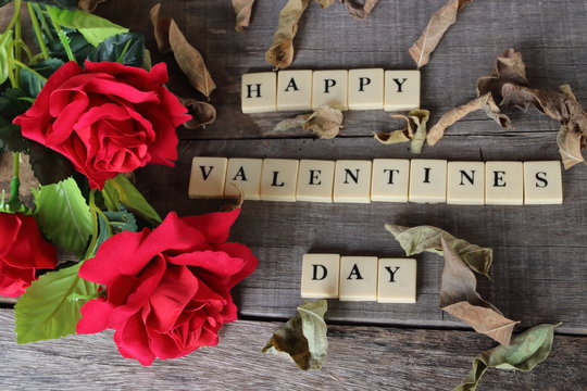 Words of Valentine's day concept by cubes of crossword game with a beautiful of artificial red roses on old wooden board background.