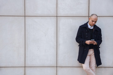 Young handsome bald businessman holding a smart phone, looking down tapping screen leaning on a white wall - technology, business, work concept 