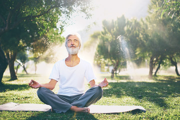 Yoga at park. Senior bearded man in lotus pose sitting on green grass. Concept of calm and...