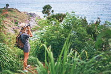 Photography and travel. Young woman in hat with rucksack taking picture on her camera enjoying sea view.
