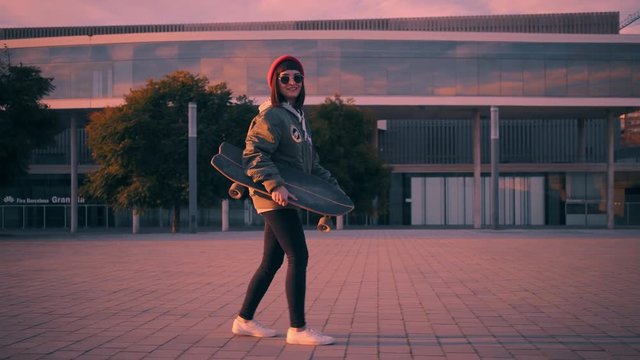 Attractive brunette weared bomber jacket,red fisherman beanie hat and tight black jeans holds small skateboard in hand and walks backward, smiling Sunset and twilight time with pastel pink lightning