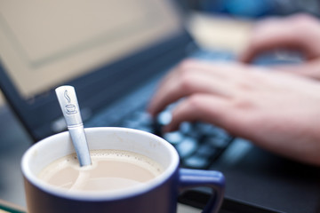 Cup of coffee with a teaspoon stamped with a coffee logo in focus with a man working on a laptop in...