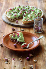 Wild Strawberry Pistachio Tart (piece) decorated with dried rose buds on wooden round board, on wooden table.